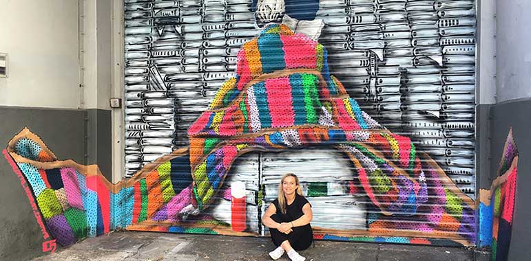 Artist setting in front of books, multi-coloured knitted blanket, coffee flask.