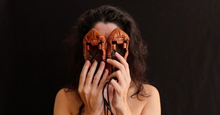 Image of the artist against a stark black backdrop, obscuring her face with a pair of brown leather albarkas shoes.