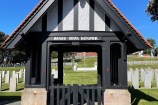 The front side of a small wooden gate building, with a picket fence and a sharp roof.