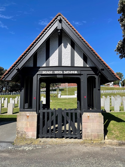 The front side of a small wooden gate building, with a picket fence and a sharp roof.