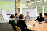 Six people sitting around a table with whiteboards in the background and a screen projector.