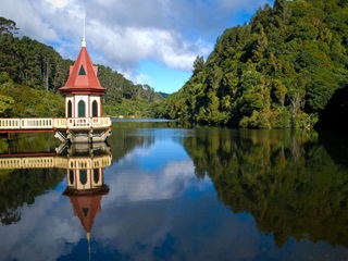 Picture of a bridge and a building over a lake, with bush in the background.