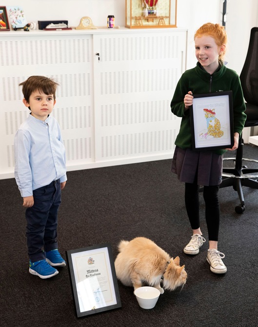 Mittens the cat sits in front of two children, one of them holding a coloured-in illustration of the cat.