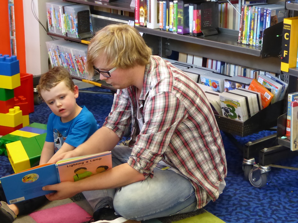 Librarian Stephen Clothier sitting on a mat reading a story book to a toddler.