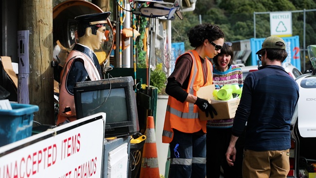 A staff member at the Tip Shop collecting a box of donations from a man and a woman, with a quirky sailor life-sized model as well as other random items in shot.