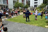 Many families sitting and standing outside in the Newtown Park Apartments complex, looking on as people dance in front of a volleyball net on grass on a sunny day.
