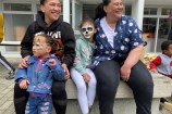Two women with big smiles, sitting on a bench, with two young children who have painted faces.