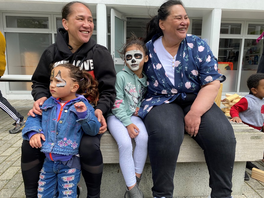 Two women with big smiles, sitting on a bench, with two young children who have painted faces.