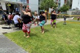 Three women in Cook Island dress dancing on the grass in front of a volleyball net, with men on seats playing guitars and apartment block in background.