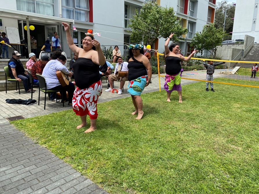 Three women in Cook Island dress dancing on the grass in front of a volleyball net, with men on seats playing guitars and apartment block in background.