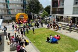 A high angle shot of the Newtown Apartment complex on a sunny day with families outside playing volleyball and on a bouncy castle, sitting on the grass, and enjoying music and dance.