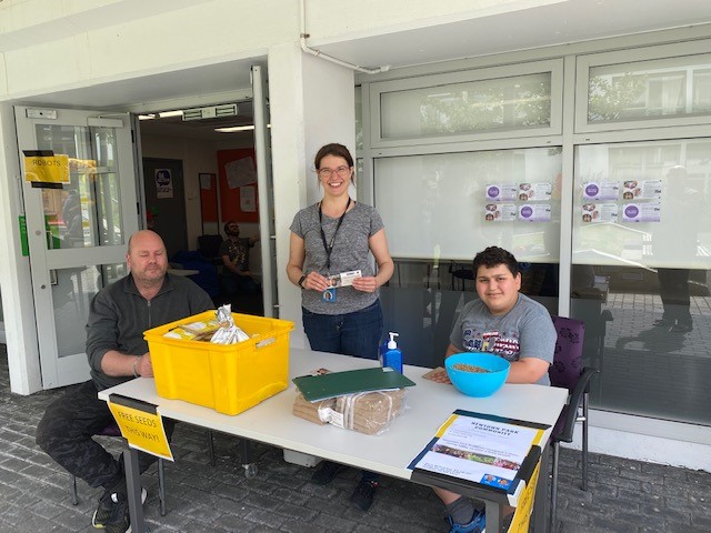 Wellington City Council's Jamie Shackleton smiling and standing in between a man and a boy, who are sitting at a table, offering free garden seeds. 