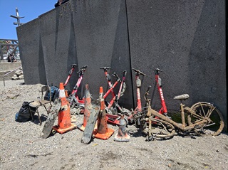 Four dirty road cones, a rusty bike, some skateboards and scooters that are a little worse for wear and rubbish that has been retrieved from Wellington Harbour, in a pile on the sand.