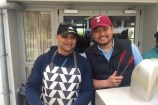 Two smiling men, in caps and aprons ,holding tongs next to a barbecue.