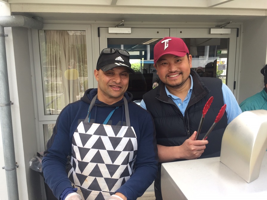 Two smiling men, in caps and aprons ,holding tongs next to a barbecue.