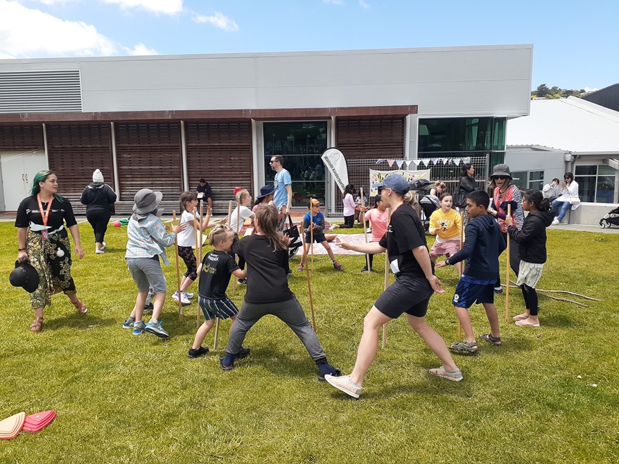 Children and adults on a grassy field enjoying the rakau challenge with sticks at the Waitohi first birthday community day.