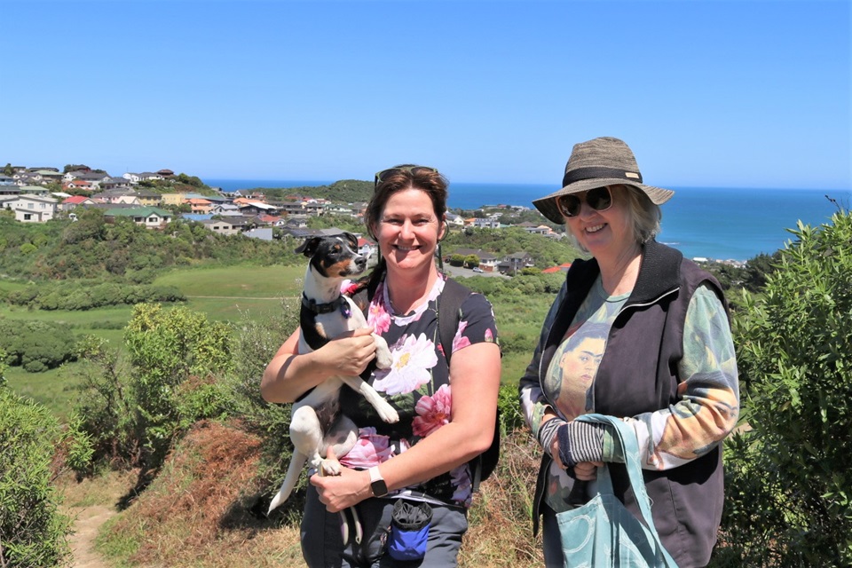 Annie Yeates, holding her Fox Terrier, and Jenny Hartley standing in the sun on the top of Tawatawa Bush Track in Owhiro Bay, with housing and the ocean in the background. 