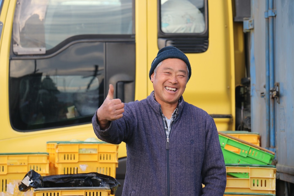 Tommy Young, from the waist up, giving a thumbs up and a big smile to the camera, in front of produce crates and the yellow cab of a truck. 