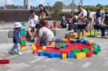 Children playing with a Lego circle outdoors on the concrete, with parents supervising from a wooden bench.
