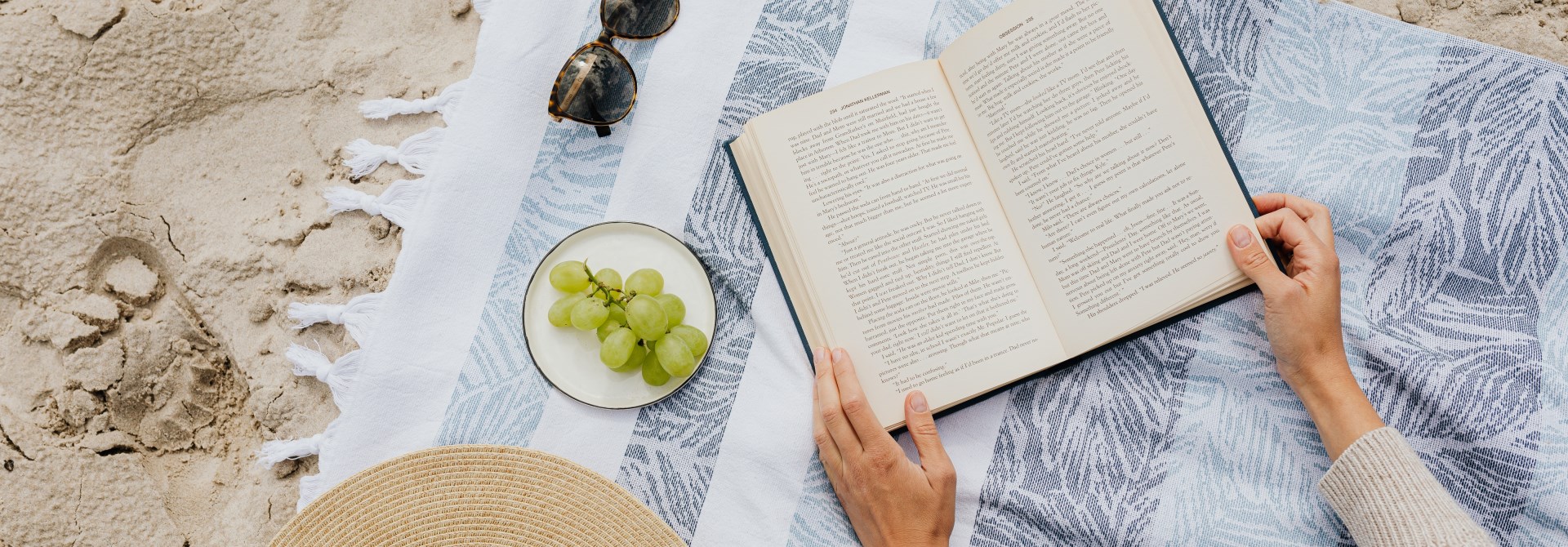 A birds-eye view of a blue and white striped towel on the sand, with a hat, some sunglasses, a drink, and a book.