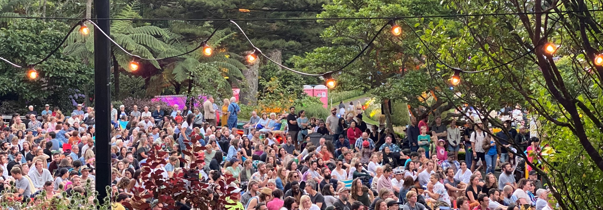 Looking through fairy lights down at a large crowd of people sitting on the lawn at the Botanic Garden summer festival, Gardens Magic, surrounded by flowers and trees.