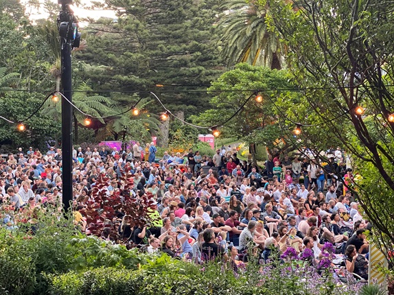 Looking through fairy lights down at a large crowd of people sitting on the lawn at the Botanic Garden summer festival, Gardens Magic, surrounded by flowers and trees.