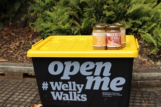 Four jars of Fix and Fogg nut butter sitting on top of the yellow and green plastic Welly Walks box, on a wooden path in amongst bush.