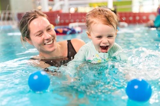 A woman in a swimsuit holding her young child as they smile and play with two blue balls in the pool.