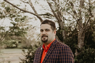 Karepa Wall in a bright red shirt with a smart check suit, pictured from the chest up, standing in front of a tree and a field.