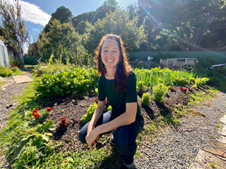 Elza, from the Seeds to Feeds initiative, smiling and crouching in a large vege patch, on a very sunny day.