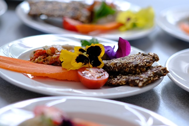 A white plate of beautifully presented food, with fresh tomato, carrot, seed bread and edible flowers.