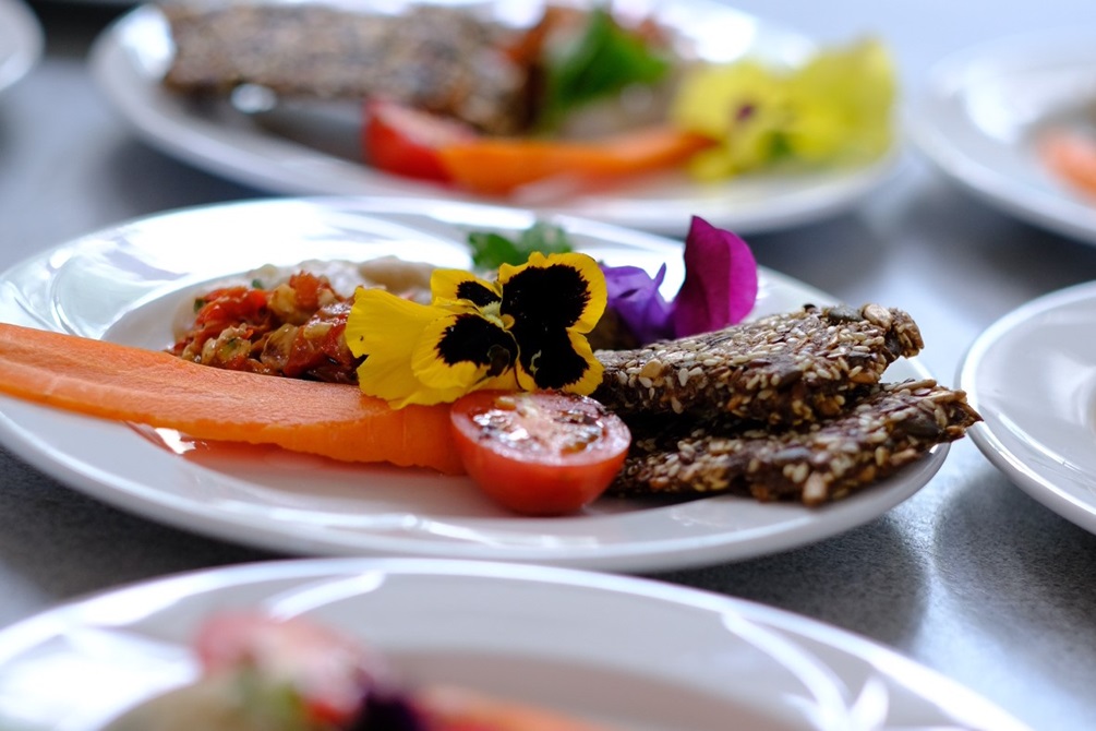 A white plate of beautifully presented food, with fresh tomato, carrot, seed bread and edible flowers.