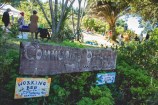 A wooden Community Orchard sign, with the gardens and people talking in the sun among trees in the background.