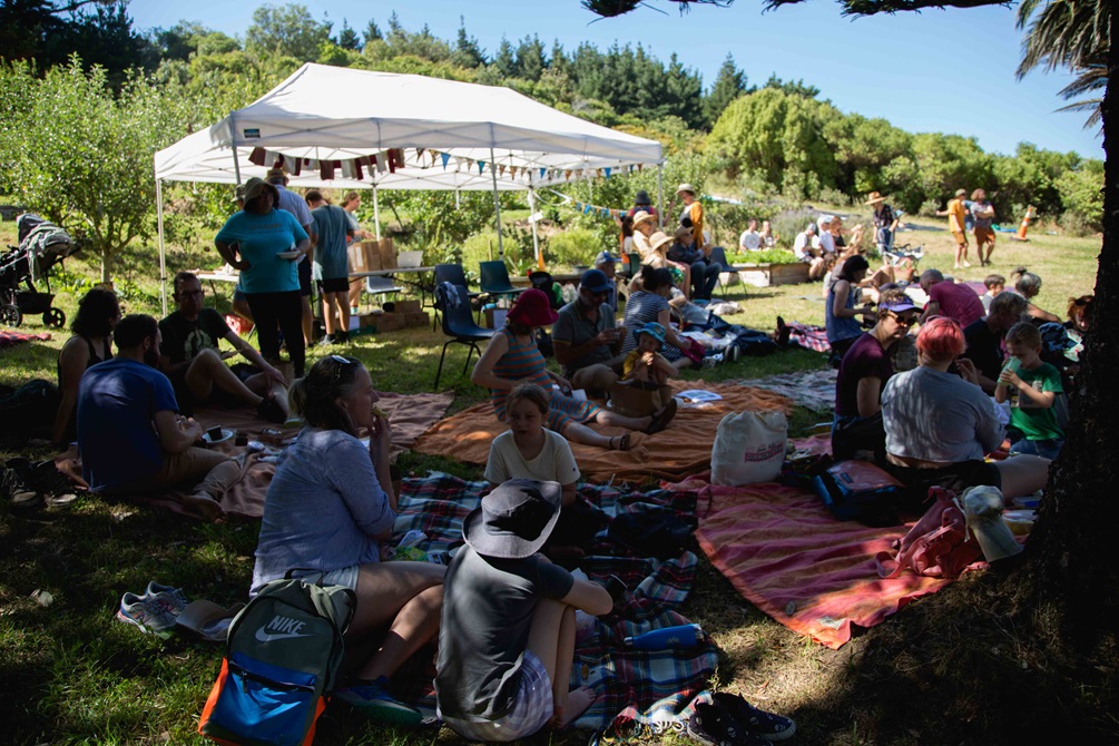 Lots of people sitting in the shade on picnic blankets, with a marquee and an orchard of trees in the sunny background.