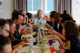 A long table with at least 15 people sitting at it drinking and eating a meal indoors with fairy lights hanging overhead.