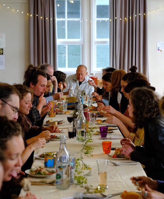 A long table with at least 15 people sitting at it drinking and eating a meal indoors with fairy lights hanging overhead.