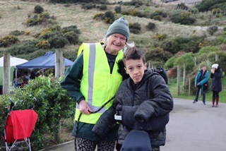 Nanny Sue in her high-vis with Cyrus Dahl on his mobility bike with people and a hill in the background.