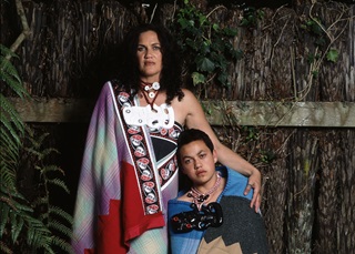 Suzanne Tamaki and her son, Rameka, in traditional Maori dress, leaning back against a punga fence.