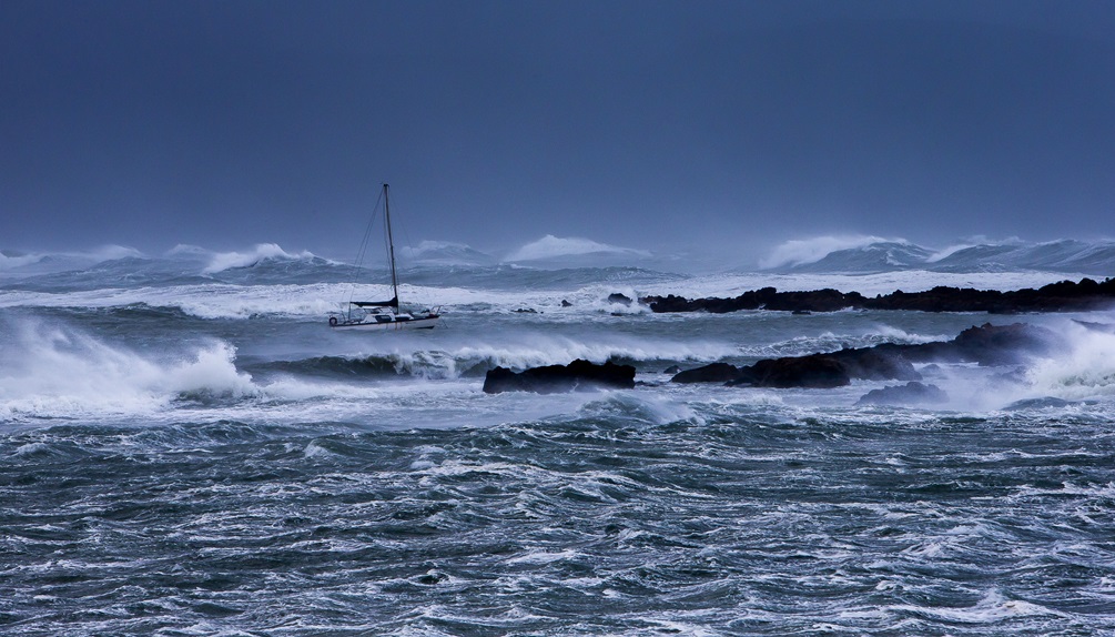 A very blue photo of severely rough seas, with waves crashing against rocks, and a small white yacht making its way through the extremely high swell.