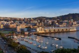 A shot of Oriental Bay on left and the boats moored and city buildings and hills behind, with a bright blue sky above.