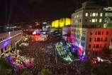 A high-angle view of a very crowded Civic Square at night time, with colourful projections on the CAB and city gallery buildings.