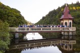 A crowd of dozens of people who have worked at Wellington City Council for 20 years or more, all standing on the cream and maroon-coloured bridge and lake lookout at Zealandia, with the structure and people reflected in the water below. Picture taken in early 2000s.