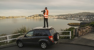 Photographer Neil Price in an orange high vis holding camera while standing on the roof of a car, looking out over Wellington harbour, with Eastbourne, Oriental Bay, and the CBD beyond.