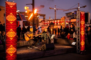 A photograph of a man and a woman singing and playing the guitar on a stage illuminated with orange light and framed by two light boxes with Māori designs on them.