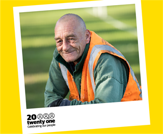 Dave Jackson, Wellington City Council's long-time lawn mower and sportsground caretaker, pictured waist up in a green jersey and orange high-vis vest, smiling, shaved head, with blurred green sportsground behind.
