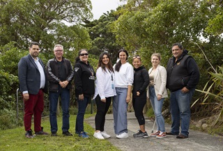 Eight people, with Karepa on the left and Kura on the right, lined up smiling on a lawn with a concrete path and native bush behind.