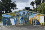 A wide colourful mural depicting people diving off a wharf and volunteers weeding in scenic environments, painted on a brick wall of a small building, with a concrete path and picnic table to the left and tall trees beyond.