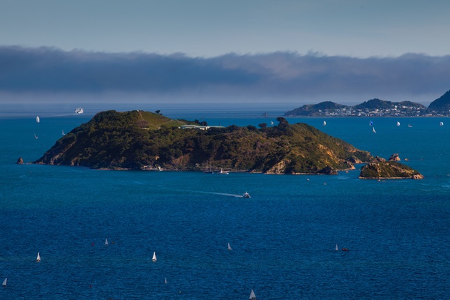 A view looking down at Somes Island in the middle of  abright blue Wellington harbour, with mist and hills beyond.