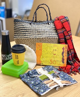 A photograph of a collection of items on a table including a whicker basket, a red tartan scarf, a black waterbottle, a yellow reusable cup, a green tupperware container, a $50 Tip Shop voucher, a material food bag and a bamboo cutlery set.