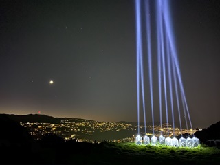 A photograph taken at night from on top of a hill with 9 lights beaming into the dark sky while the street lights of the city glitter gold in the foreground.